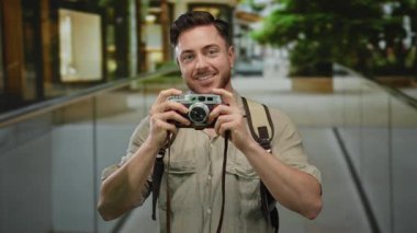 Sakallı genç bir adam şehir manzarasını bir turist olarak yakalıyor, kamera elinde, gülümsüyor ve açık bir sokakta sırt çantası takıyor..