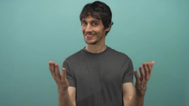 Gri tişörtlü gülümseyen genç adam elleri açık bir deniz mavisi stüdyo arkaplanına işaret ediyor;.