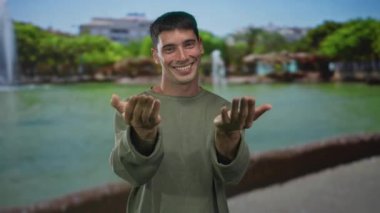 Yeşil tişörtlü adam sokakta elleri ile çağıran, güneşli gökyüzü altında geniş bir gülümseme; dostluk sıcaklığı davet.