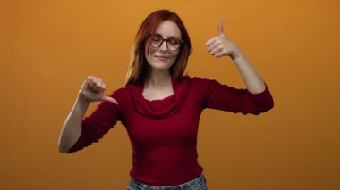 Turuncu arka planda baş parmak hareketi yapan bir kadın kırmızı kazakla karışık duygular ifade ediyor ve gözlüklü bir kadın hem onaylıyor hem de onaylamıyor..