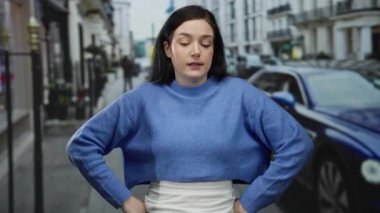 Mavi kazaklı kadın, arka planda arabaları ve binaları olan şehir caddesinde rahat bir şekilde duruyor, hareketli bir şehir ortamında sakin tavırlar sergiliyor..