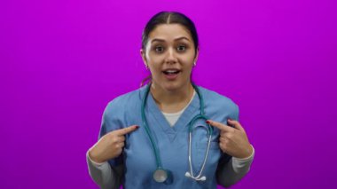 Mavi önlüklü, steteskoplu İspanyol hemşire, kendini izole edilmiş pembe duvar arka planına işaret ederek şaşırmış görünüyor..