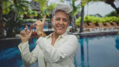 Etrafta ahşap saat, palmiye ağaçları ve şezlonglar olan bir kadın, havuz kenarını işaret ediyor..