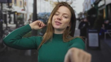 Yeşil kazaklı genç beyaz kadın parmaklarını objektife uzatırken gülümsüyor arkasında da bulanık bisikletler var..