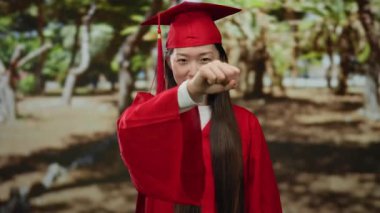 Parktaki mezuniyet kutlamasında kırmızı şapkalı ve cüppeli genç bir kadın uzanmış yumruğuyla başarıyı ve başarıyı simgeliyor..