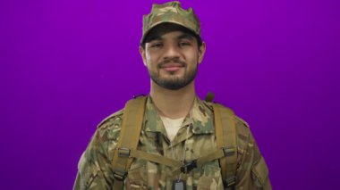 Kamuflaj üniformalı, künyeli, sırt çantalı İspanyol bir adam stüdyoda kollarını kavuşturuyor..