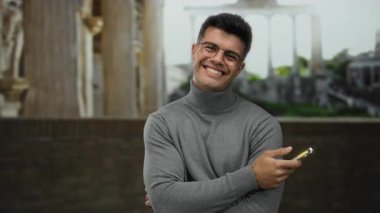 Arkaplanda Roma harabeleri olan akıllı telefona gülümseyen genç bir adam tarihi açık hava ortamında modern teknoloji kontrastı sergiliyor..