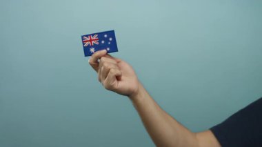 Avustralya bayrağını mavi bir duvara dayamış beyaz bir adam, ulusal gurur ve kimliği basit ve izole bir ortamda vurguluyor..