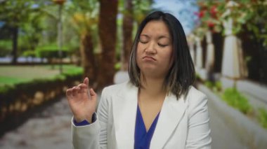 Parkta beyaz ceket ve mavi bluz giyen genç bir kadın elini kullanarak yeşillik ve patikanın arka planına karşı olduğunu gösteriyor..