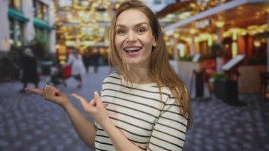 Çizgili bluzlu genç kadın gülümsüyor ve meşgul caddede parlayan kafenin ışıkları altında parmak ucuyla işaret ediyor; heyecan.