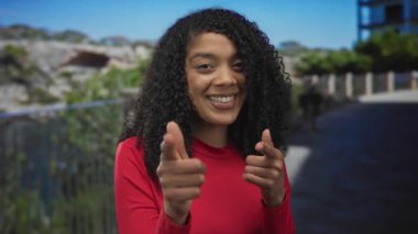 Kırmızı gömlekli kadın, güneşli parkta kaldırım kenarında çitin yanındaki kamerayı işaret ediyor..