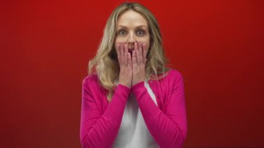 Genç bir kadın heyecanlı bir şekilde kırmızı bir arkaplanda hareketler yapıyor, sarı saçları pembe bir kazakla çelişiyor, anlamlı bir sürpriz ve neşe anı yakalıyor..