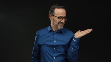 Mavi gömlekli İspanyol bir adam, avucunun içi açık bir şekilde siyah arka plana karşı işaret ediyor. Kendine güvenen bir ifade takınıyor ya da sunuyor.