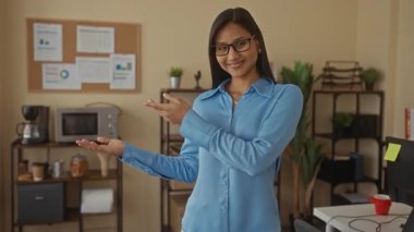 Mavi gömlekli genç bir kadın arka plandaki mantar panoda bitkiler, mikrodalga fırın ve çizelgelerle modern ofis ortamında gülümsüyor ve el kol hareketi yapıyor..