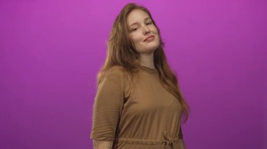 Kahverengi elbiseli genç beyaz kadın canlı mor stüdyo arkaplanına karşı göğsünü işaret ediyor..