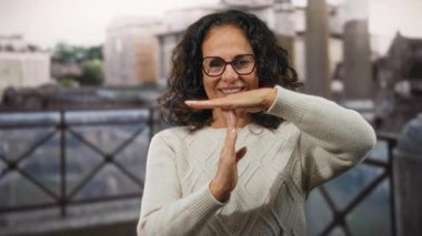 Kıvırcık saçlı, olgun, gülümseyen bir kadın şehir ortamında, beyaz bir süveter giyerek mola veriyor..