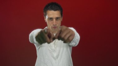 Beyaz tişörtlü genç İspanyol, stüdyoda kırmızı duvara karşı parmak ve kamerayla işaret ediyor..