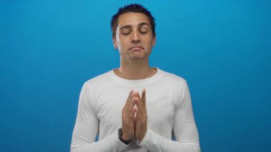 İzole edilmiş mavi arka plan üzerinde beyaz tişörtlü genç İspanyol adam ellerini meditasyona sıkıştırıp rahatladığını ve iç huzurunu göstererek huzurlu görünüyor.
