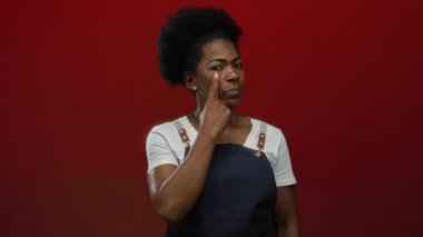 Önlüklü Afrikalı kadın, izole edilmiş kırmızı arka planda parmaklarıyla göze dokunuyor, düşünceli ve içgözlem yapan önemli görev ve kararları değerlendiriyor..