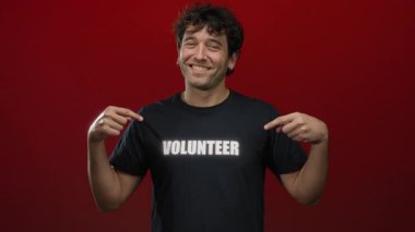 İspanyol adam gülümsüyor ve canlı kırmızı bir arkaplan karşısında siyah tişört üzerindeki gönüllü metne işaret ediyor..