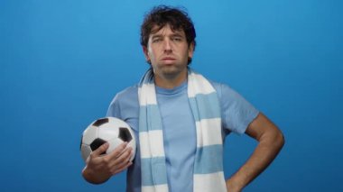 İspanyol adam futbol topunu tutuyor mavi atkı takıyor ve mavi arka planda spor hevesini tasvir eden kararlı bir ifade sergiliyor..