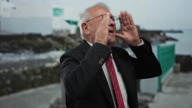 Takım elbiseli kıdemli adam deniz kenarına bağırıyor, okyanus arka planında, acil durum ya da acil durum temasını aktarıyor..