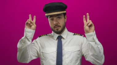 Pilot üniforması içinde sakallı genç adam pembe arka plana karşı jest yapıyor, şakacı bir şekilde alaycılık ve mizah yapıyor..