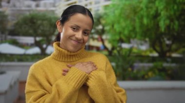 Sarı süveter giyen genç İspanyol kadın şehir caddesinde çevrenin yeşillik ve binalarla çevrili olduğu yerde içten içe gülümsüyor. Mutluluk ve memnuniyet saçıyor.