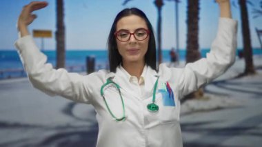 Steteskoplu kadın doktor, deniz kenarında palmiye ağaçlarıyla dolu bir yerde kalp jesti yapıyor. Üniformayla dışarıdaki sükuneti kucaklıyor..