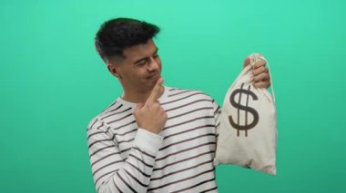 Çizgili gömlekli İspanyol adam yeşil arka planda para çantasına işaret ediyor, zenginlik ve başarı hissi veriyor..
