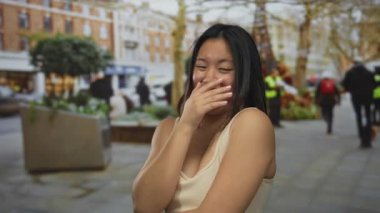 Şehir arka planında bulanık şehir arka planında, açık havada gülen bir kadın aydınlık bir günde gençlik neşesi ve günlük giysiler sergiliyor ve insanlar uzaklarda yürüyor..