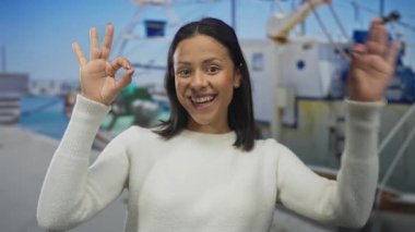 Neşeli bir şekilde gülümseyen kadın, güneşli bir günde arka planda kayıklar olan canlı bir sahil limanında iki eliyle güzel bir jest yapıyor..