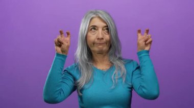 Gri saçlı bir kadın mor arka planda poz veriyor. Deniz mavisi süveter giyiyor. Konsept espri veya iğneleme yapıyor.