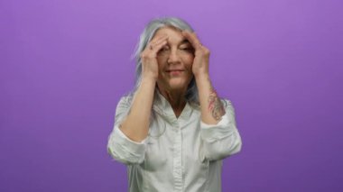 Yaşlı, gri saçlı, dövmeli bir kadın, ellerini şakaya vurarak yüzünü kapatıyor, sonra gülümsüyor, parlak mor bir arka plana karşı izole edilmiş, neşeli ve çekici bir görüntü yaratıyor..