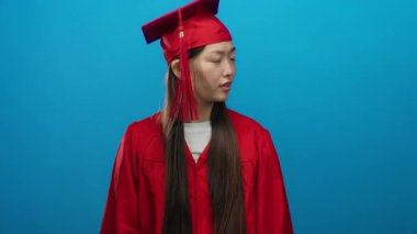 Kırmızı mezuniyet elbisesi giymiş genç bir kadın canlı mavi bir arka plana karşı poz veriyor, gururlu ve heyecanlı..