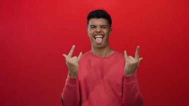 Adam şakacı el hareketi yapıyor ve dili dışarı çıkıyor, pembe gömleği kırmızı arka planda giyiyor..