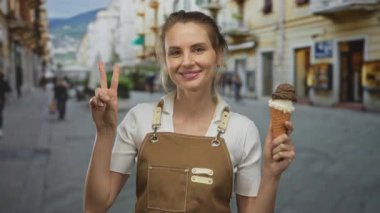 Önlüklü, elinde dondurma külahıyla gülümseyen bir kadın açık havada güneşli şehir ortamında barış işareti yapıyor..