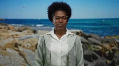 Deniz kenarında güvenle duran, arkasında kayalar olan bir kadın, açık mavi gökyüzünün altında zarif bir varlığı tasvir ediyor, açık havada gözlük takıyor..