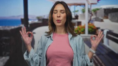 Deniz kenarında meditasyon yapan bir kadın, gözleri gevşemiş, canlı çiçekler ve okyanus manzarasıyla çevrili, sahilde huzur ve sükuneti çağrıştırıyor..