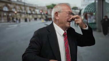 İş elbisesi giymiş yaşlı bir adam kalabalık bir sokakta dışarıda dikilirken kötü kokuya karşı burnunu sıkar..