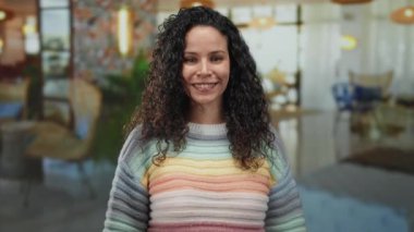 İspanyol kadın, kıvırcık saçlı, kapalı bir otel ortamında renkli çizgili bir kazak giymiş bulanık bir arka planda çeşitli düşünceli pozlar veriyor.