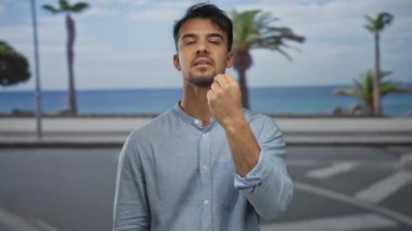 Arka planda palmiye ağaçları olan deniz kenarındaki gezinti alanında hayal kırıklığına uğramış genç İspanyol bir adam mavi bir gömlek giyiyor ve manzaralı okyanus manzarasının yanında dikiliyor..