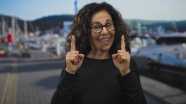 Gözlüklü neşeli kadın arka planda tekneler olan deniz manzaralı bir limanı işaret ediyor..