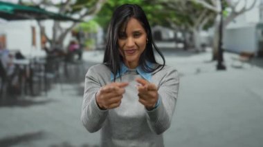 Gülümseyen bir kadın, ağaçlarla kaplı bir caddeye gelip gündüz vakti açık havaya davet ediyor, şehir ortamında günlük gri süveter giyiyor..