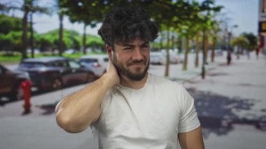 Dışarıda sakallı genç bir adam güneşli bir sokakta boynunu tutuyor. Sıradan bir şehir ortamında rahatsızlık ya da acı olduğunu gösteriyor..