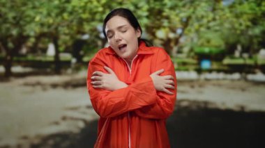 Turuncu hapishane tulumlu kadın yeşil ağaçlarla çevrili bir açık hava parkında kendini kucaklıyor, içsel gözlem ve sükunet duygularını aktarıyor..