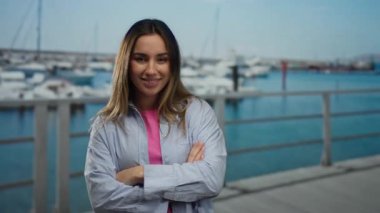 Kolları çaprazlanmış bir kadın, güneşli bir günde deniz kenarındaki marinada teknelerle ve plaj arkaplanıyla gülümsüyor, açık havada güven ve rahatlama yayıyor..