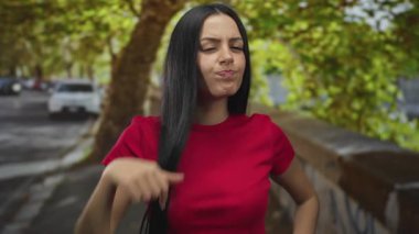 Kırmızı tişörtlü İspanyol kadın, arka planda ağaçlar ve arabalarla şehir sokaklarında öfkesini sergiliyor, güçlü duygusal ifadeyi vurguluyor..