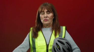 Sarı bir yelek giyen ve kırmızı bir arka plana karşı kask tutan bir kadın, bir inşaat ya da güvenlik hikayesi ima ederek şaşkınlığını dile getiriyor..