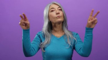 Yaşlı gri saçlı kadın, sakin bir ifadeyi gösteren izole bir mor arka plana karşı barış işareti yapıyor..